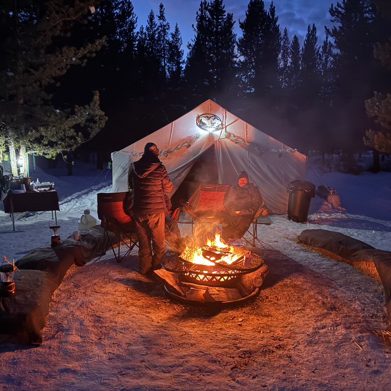 Two people around a campfire in front of a tent at night in a snowy forest.